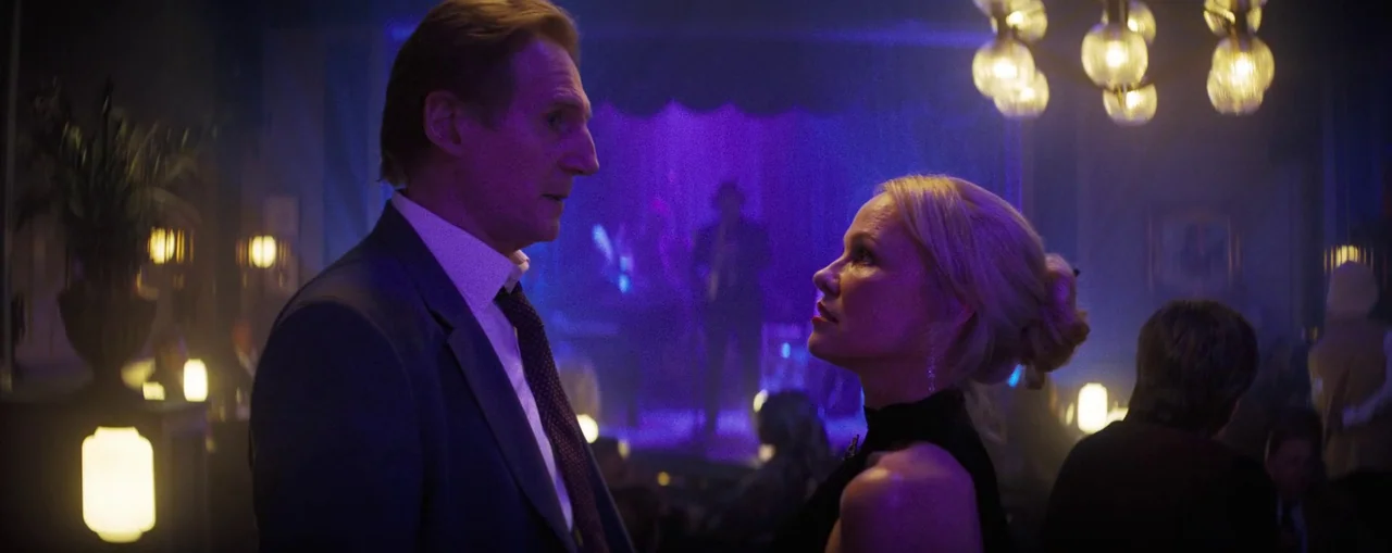 The Naked Gun (2025) movie still: medium — A dimly lit club features a purple-lit stage with performers in the background. A man and a woman stand…