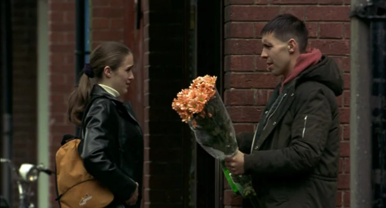 A Room For Romeo Brass (1999) movie still: medium — A man and woman stand against a weathered red brick wall during the day. The man holds out a…