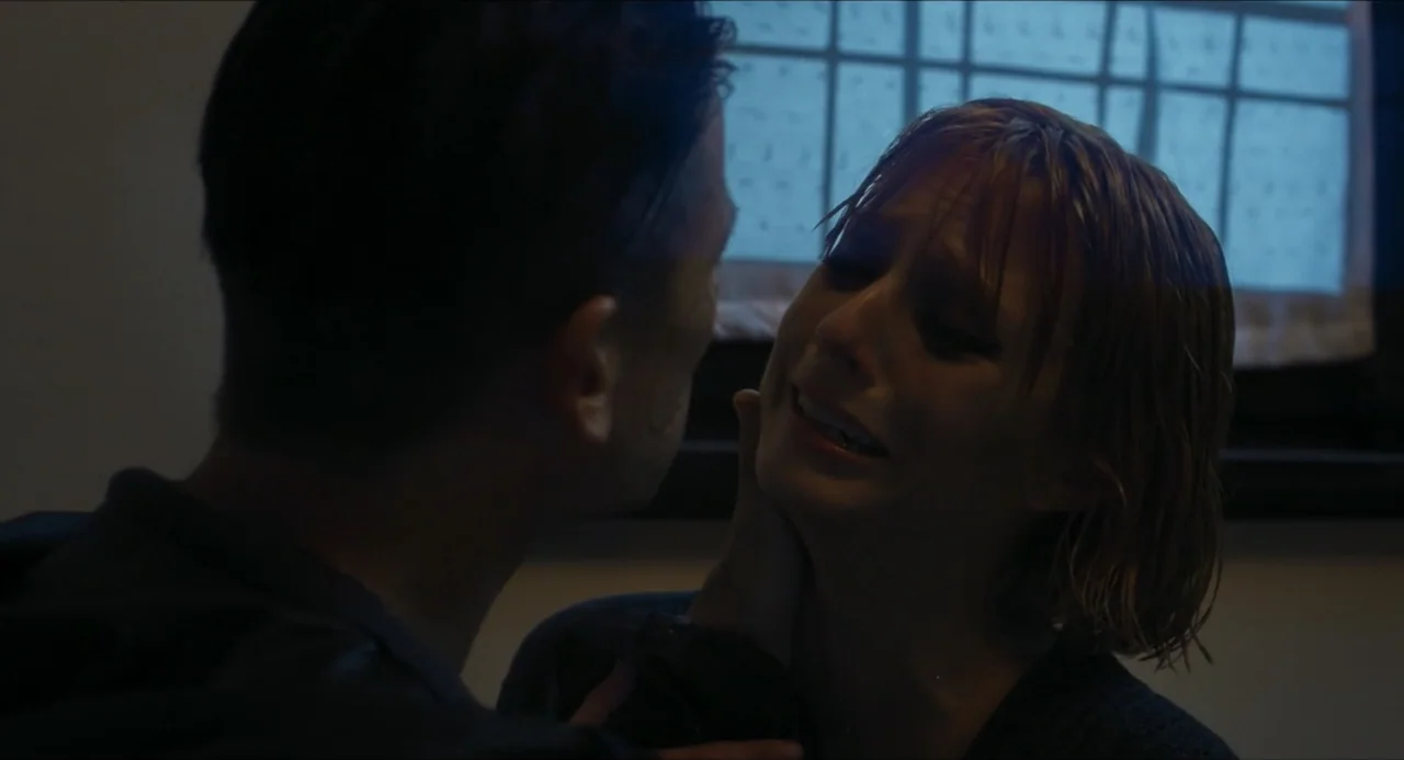 Echoes Of A Crime (2025) movie still: closeup — A dimly lit room at night features a window with horizontal panes in the background. A man gently…