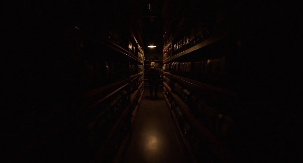 Nightmare Cinema movie still: wide — A dark aisle flanked by tall metal shelves filled with stacked storage containers. A solitary figure stands at…
