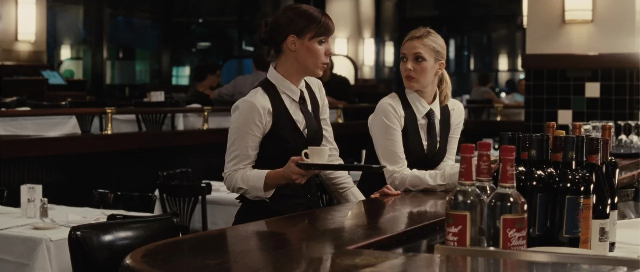 Going the Distance (2010) movie still: medium — A quiet restaurant interior with a dark wood bar and white-clothed tables. Two waitresses in…