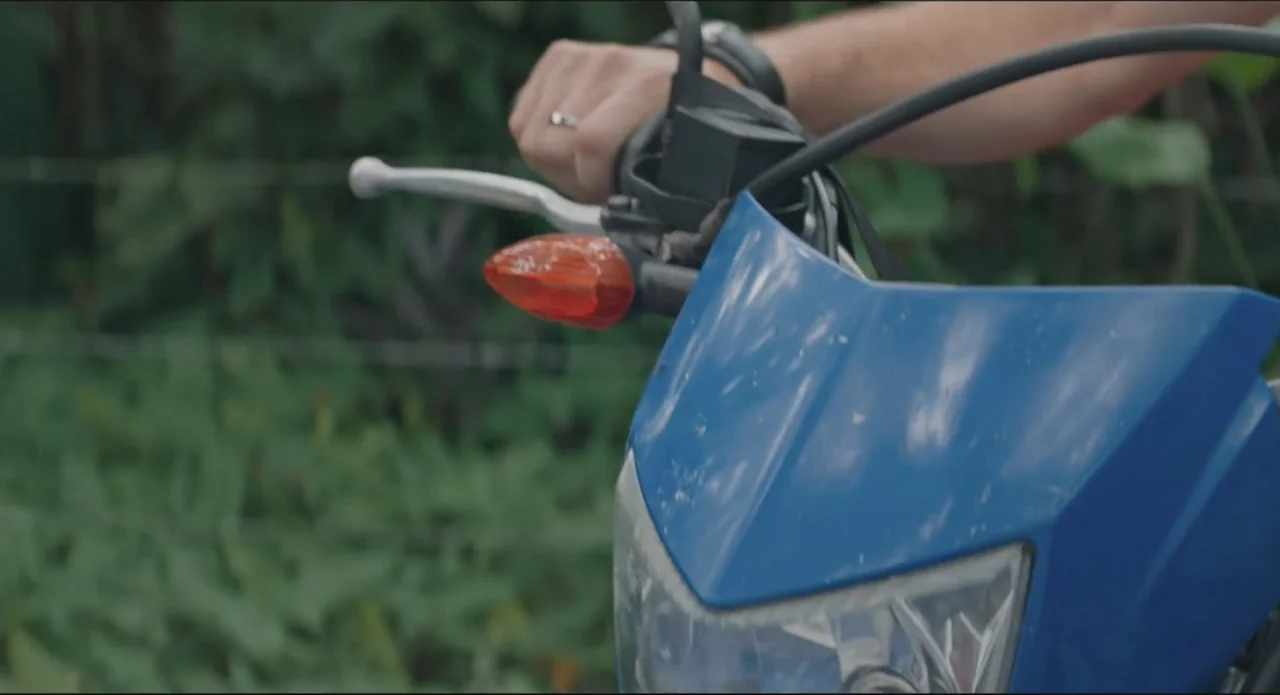 Proof of Love (2022) movie still: closeup — A close-up of a blue motorcycle handle and turn signal in a rural setting. A hand with a wedding ring…