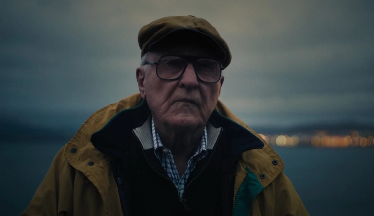 Land Ho! (2014) movie still: medium — An elderly man stands at a coastal location during the low light of dusk. Blurred bokeh city lights are…