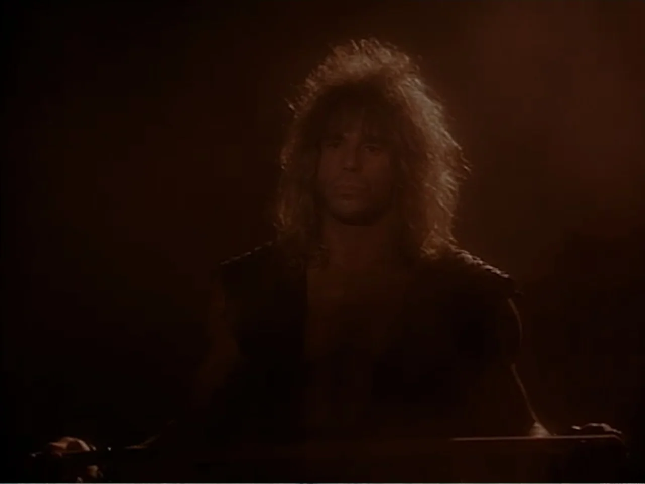 Firepower (1979) movie still: medium — A dark, indistinct interior setting with warm, low-key lighting focused on the subject. A man with long hair…