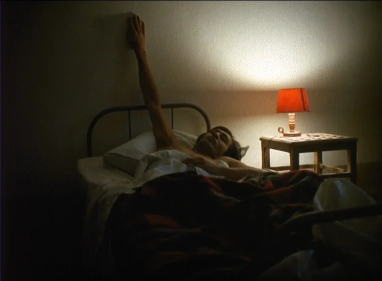 La Collectionneuse (1967) movie still: medium — A dim room features a simple iron bed frame positioned against a plain wall. A man lies in the bed…