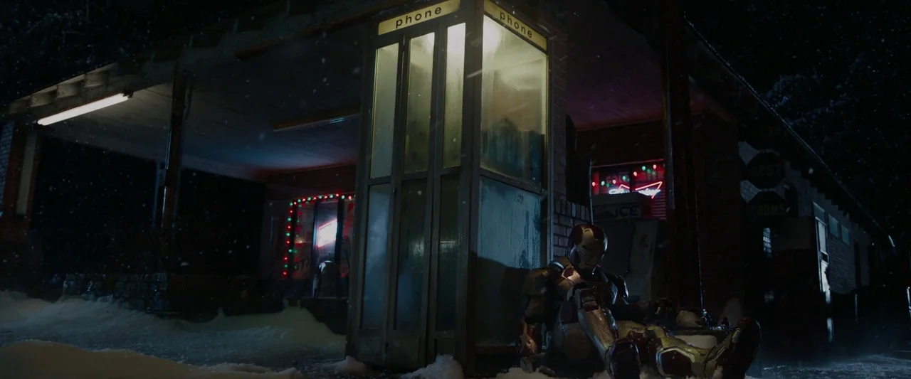Iron Man 3 (2013) movie still: wide — A dark, snow-covered gas station at night features a lone glass phone booth. A metallic armored figure in red…