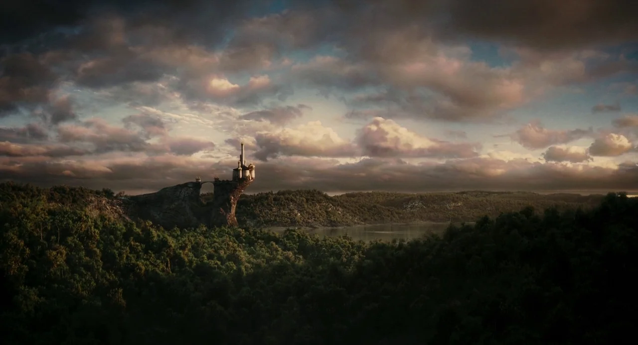 Mirror Mirror (2012) movie still: establishing — A stone castle structure sits atop a high, dark rocky cliff overlooking a lake. Dense forest covers…