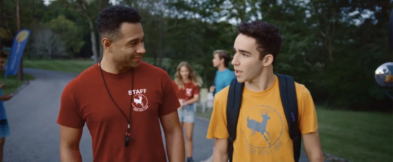 Camp Hideout (2023) movie still: medium — An outdoor paved path at a camp with green trees in the background. A staff member walks and talks with a…