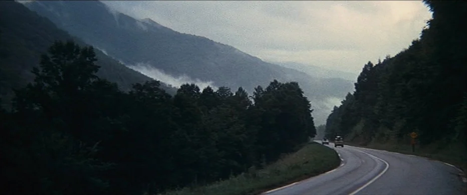 Deliverance (1972) movie still: establishing — A winding asphalt road curves through a dense forest bordered by towering misty mountains. Two small…