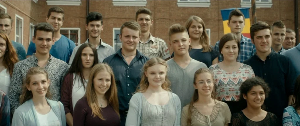 Graduation (2016) movie still: medium — A group of diverse teenagers poses for a photograph in front of a brick building. The students are arranged…