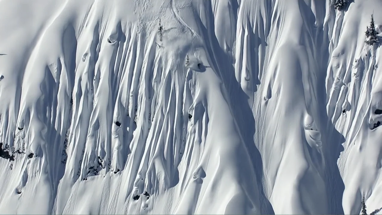 Dear Rider: The Jake Burton Story (2021) movie still: wide — A steep mountainside covered in deep snow features dramatic gullies and ridges. Sparse…