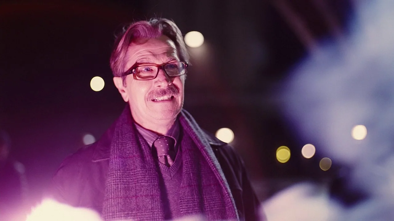 The Dark Knight Rises (2012) movie still: medium — A man stands outdoors at night in front of out-of-focus city lights. He wears glasses and a scarf…