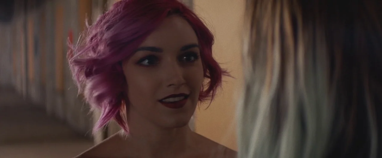 Poser (2021) movie still: closeup — A dimly lit indoor hallway with a blurry figure visible in the foreground. A woman with short pink hair faces…