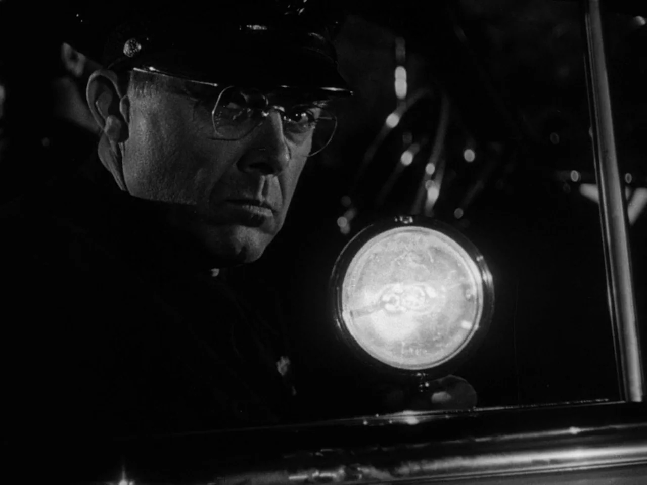 The Prowler (1951) movie still: closeup — A dark, cramped interior of a vintage vehicle at night. A police officer holds a glowing flashlight…