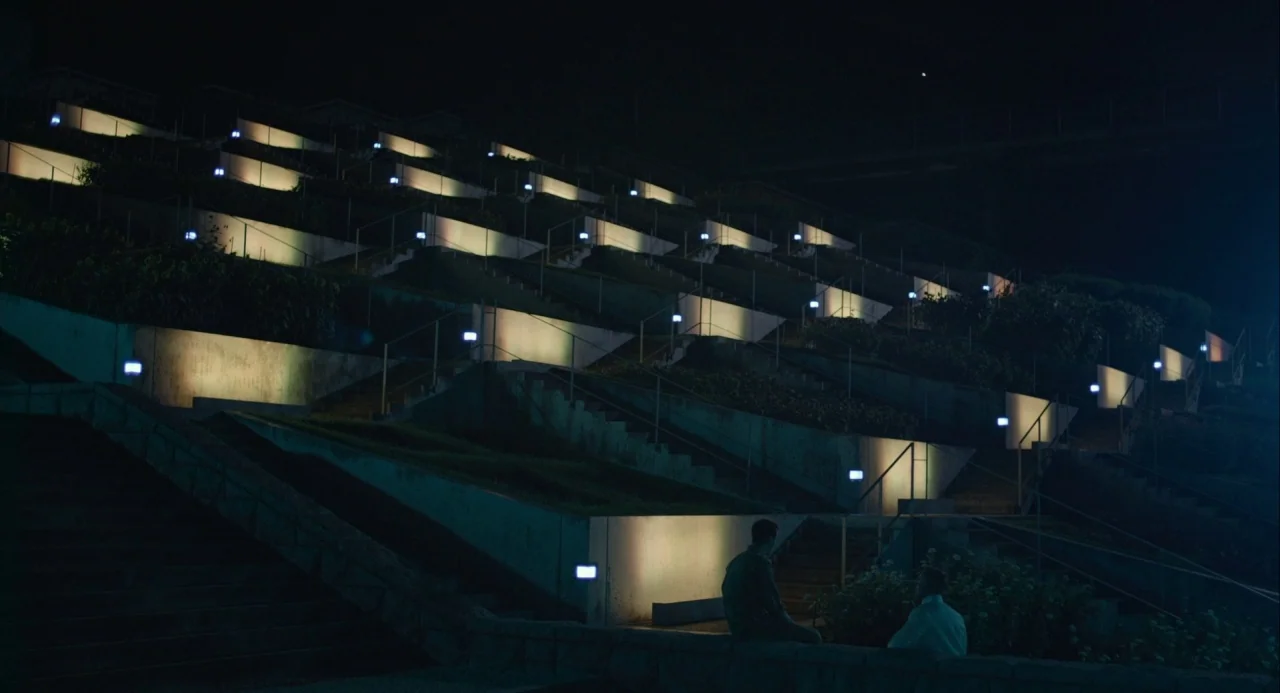 Equals (2015) movie still: wide — A multi-tiered outdoor space features concrete retaining walls and staircases at night. Two men sit on a low ledge…