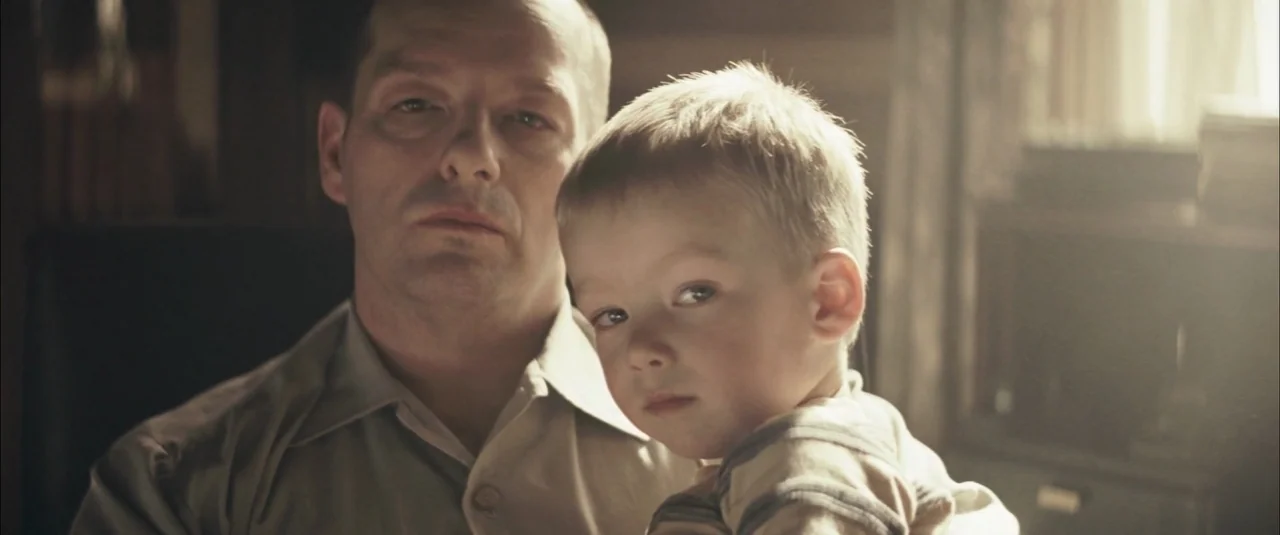 The Grey (2012) movie still: closeup — A dimly lit domestic interior space features a man and child. The man holds the child securely while they…