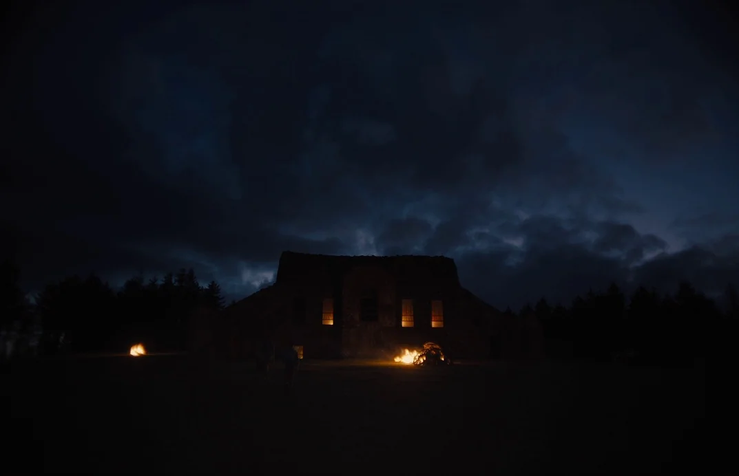 Gretel & Hansel (2020) movie still: wide — A stone structure sits under a dark, cloudy night sky in an open field. Two small fires burn in front of…