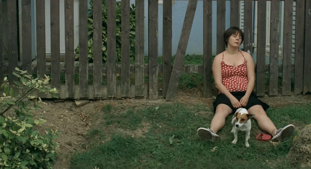 Water Lillies (2007) movie still: fullbody — A woman sits on the grass against a weathered wooden fence in a yard. She rests with her eyes closed…