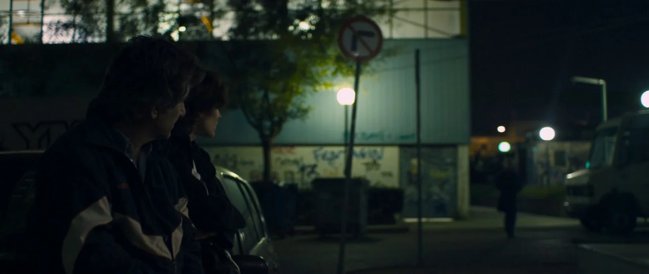 Alps (2011) movie still: medium — An urban parking lot at night with graffiti-covered walls and a distant streetlamp glow. Two people lean against a…