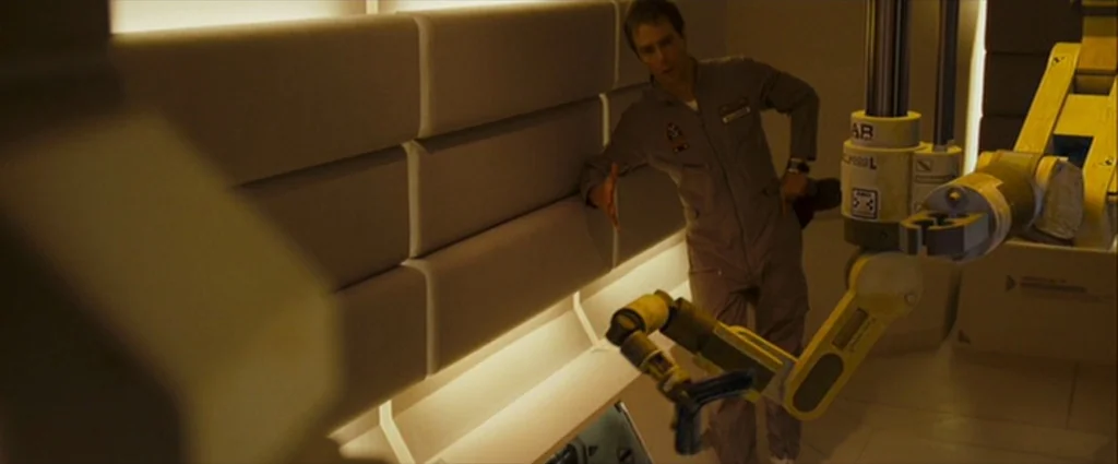 Moon (2009) movie still: medium — A minimalist space with light beige upholstered wall panels and yellow mechanical fixtures. A man in a jumpsuit…