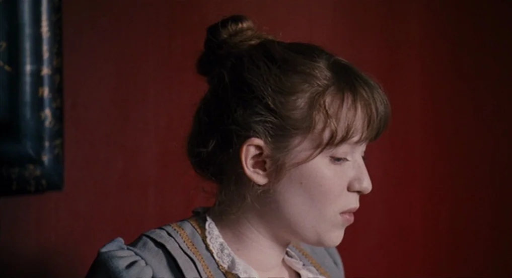 House of Tolerance (2011) movie still: closeup — A woman stands against a deep red wall in an interior setting. She has her hair in a high bun and…