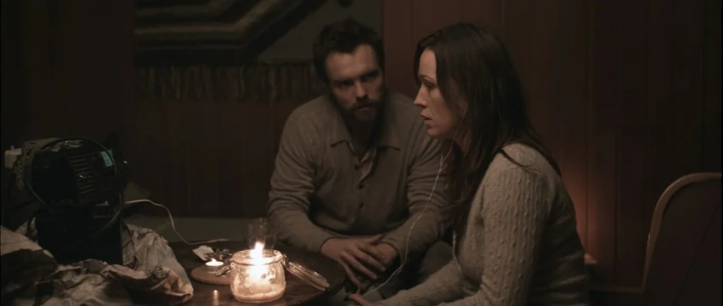 One Hundred Mornings (2009) movie still: medium — A dimly lit room features wood-paneled walls and a woven tapestry in the background. A man and a…