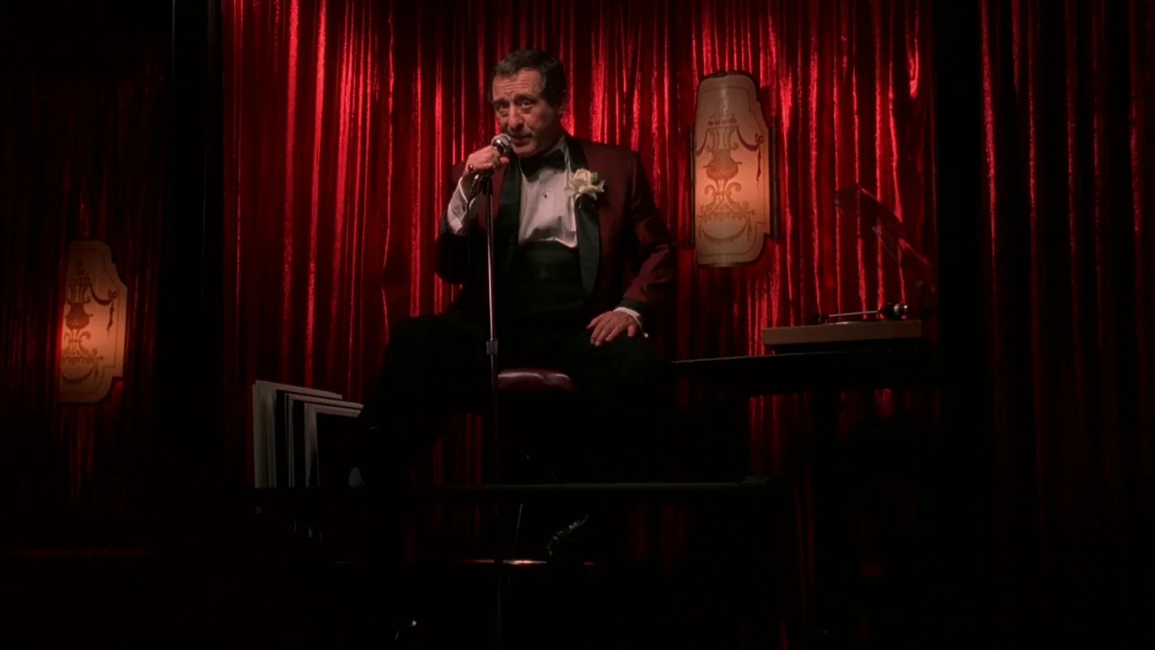 Auto Focus movie still: medium — A dark lounge features floor-to-ceiling red velvet curtains and dim wall-mounted decorative lamps. An elderly man…