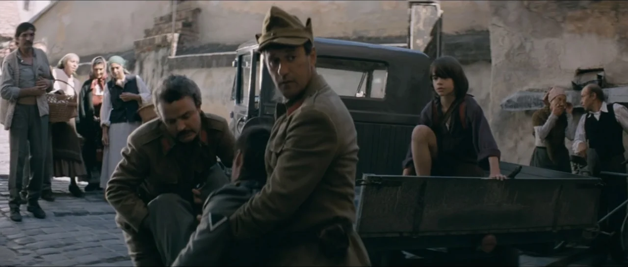 A Farewell to Fools (2013) movie still: medium — A narrow stone street features a weathered cargo truck parked against textured stone walls. Two…