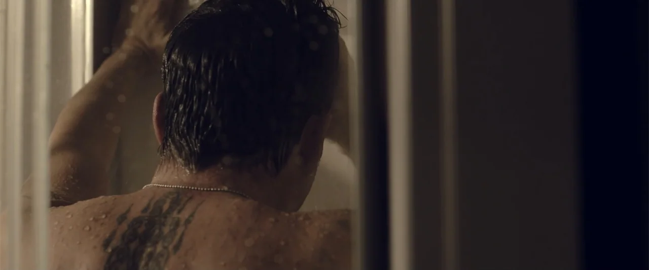 About Us (2017) movie still: closeup — A dark tiled shower stall contains a man under the stream of water. The subject faces the wall with his hand…