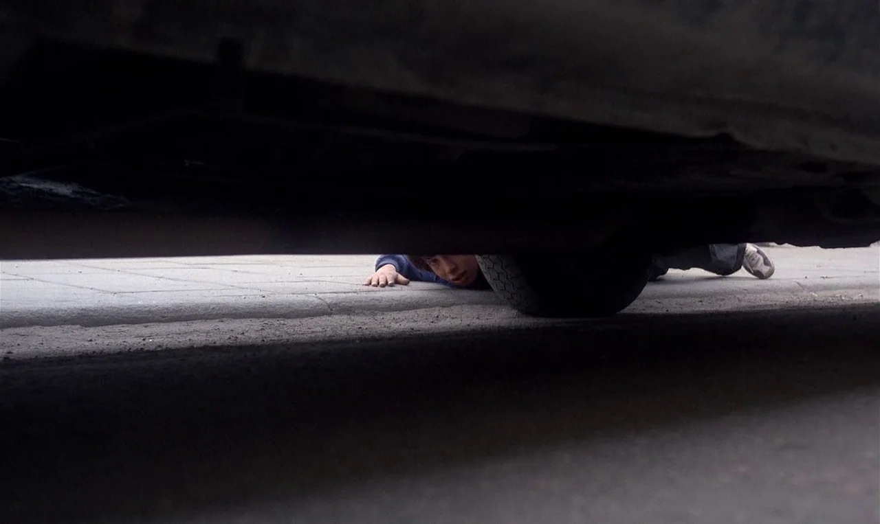 Man on the Roof (1976) movie still: wide — A view from beneath the chassis of a parked vehicle onto paved ground. A young person lies face-down on…