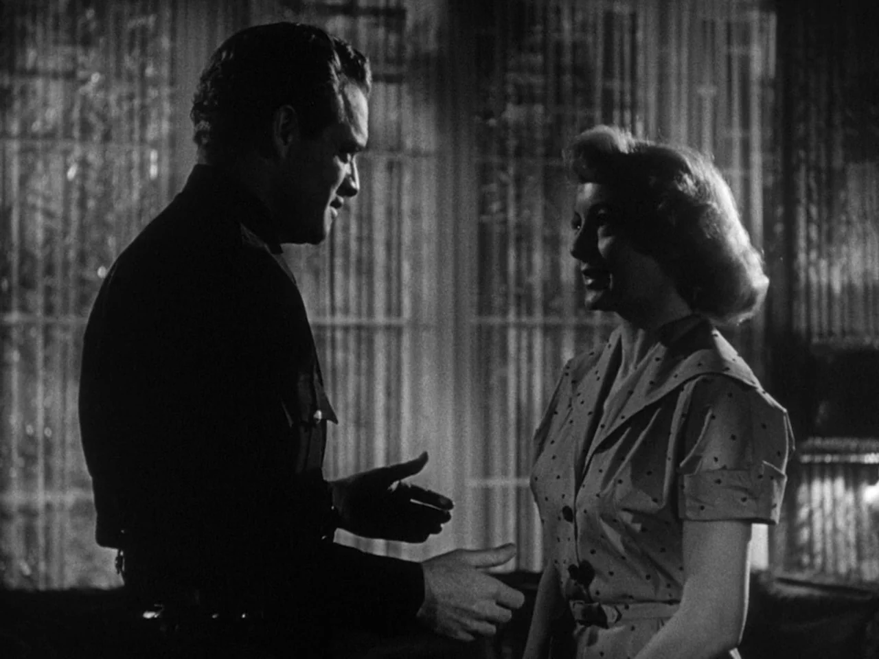 The Prowler (1951) movie still: medium — A dark indoor space features thin vertical curtains partially obscuring the light source behind. A man and…