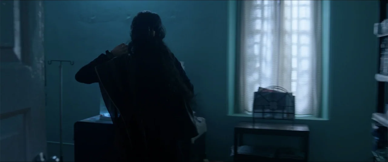 Cheekatilo (2026) movie still: medium — A dimly lit room with blue walls and a window covered by a curtain. A person with long hair stands in the…