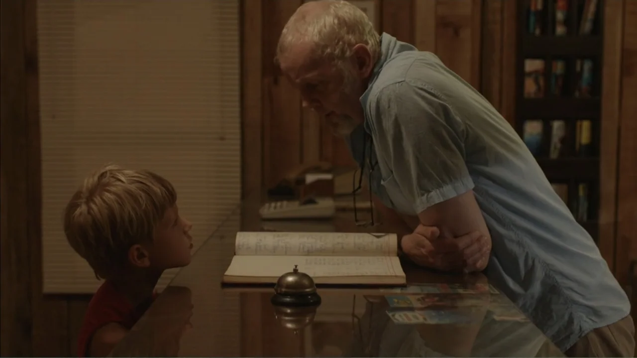 Boy (2010) movie still: medium — A wooden hotel front desk features a guest register and a small brass bell. An elderly man in a light blue shirt…