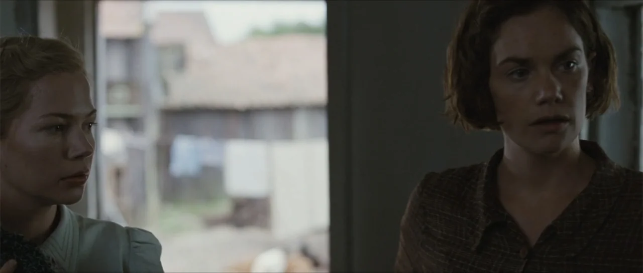 Suite Française (2015) movie still: medium — A rustic building interior looks out toward a rural courtyard with blurred structures. Two women stand…