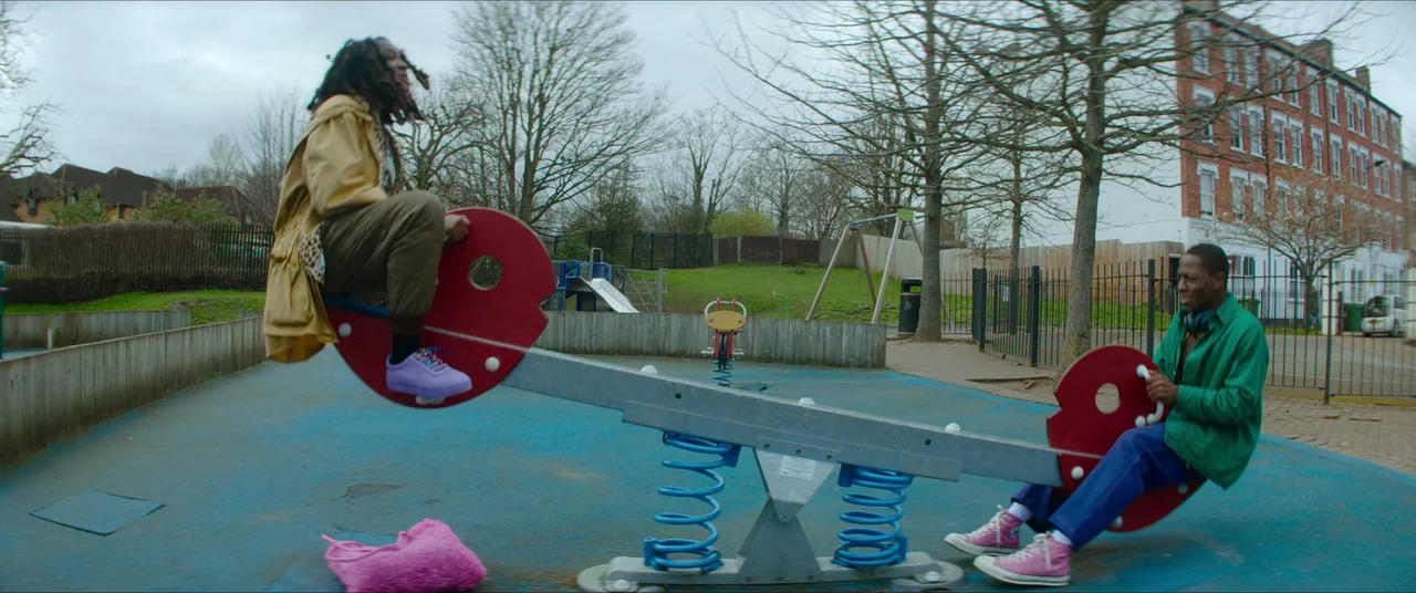 Rye Lane (2023) movie still: wide — An urban playground with a blue rubber floor and metal seesaw equipment. Two young adults balance each other…