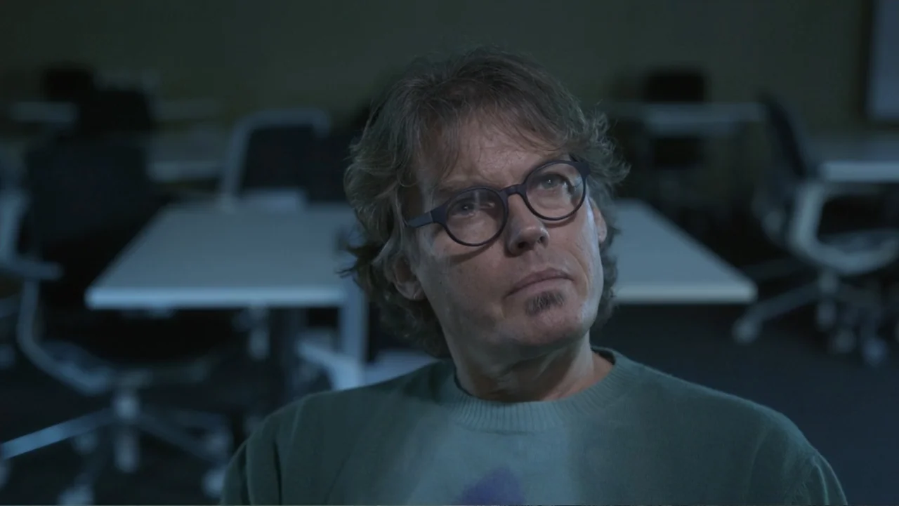 Predators (2010) movie still: closeup — A dimly lit modern workspace features white tables and blurred chairs in the background. A middle-aged man…