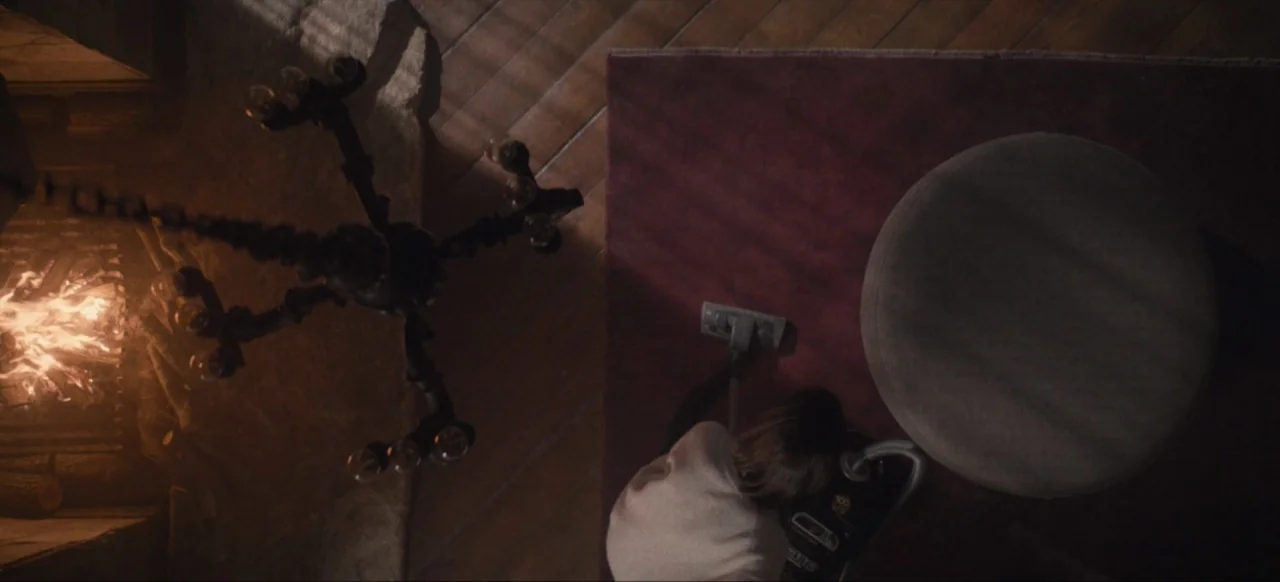 Housewife (2017) movie still: wide — A top-down view of a dark room with wooden floors and a large fireplace. A woman stands on a red rug vacuuming…