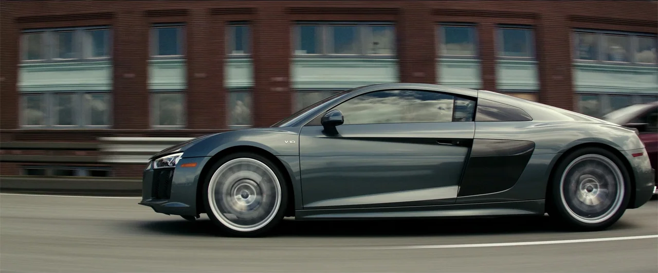 Fifty Shades Freed (2018) movie still: medium — A grey sports car travels at high speed on a multi-lane highway. The blurry brick building…