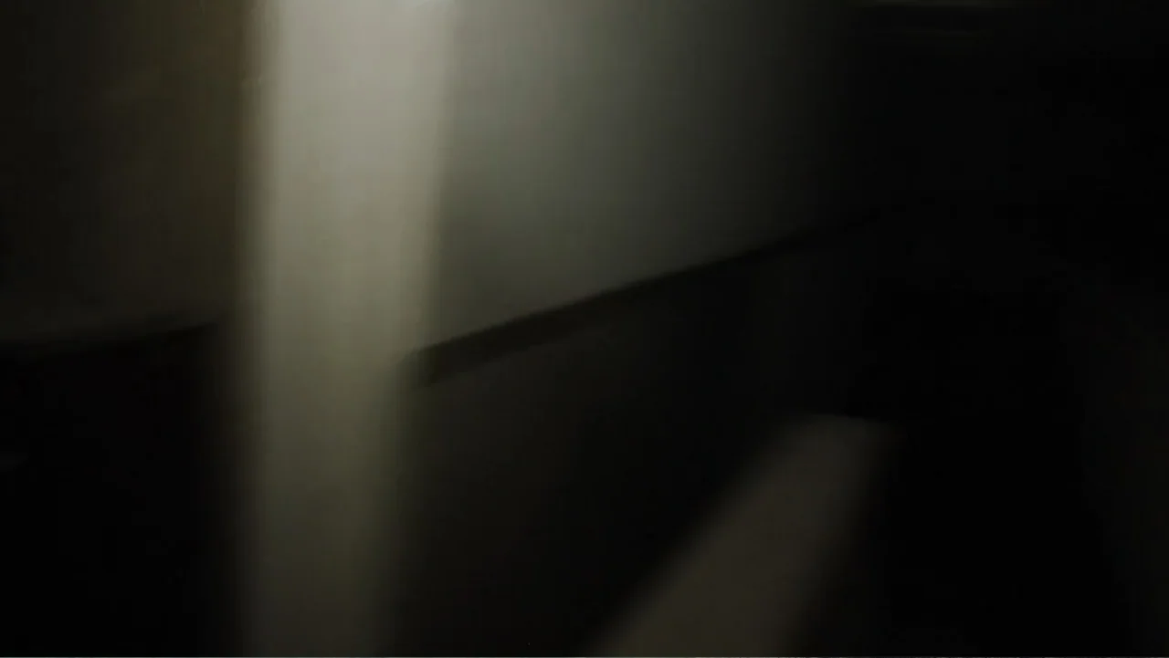 Lost (2010) movie still: establishing — A dark interior space features a blank wall intersected by a singular light source. The dimly lit…