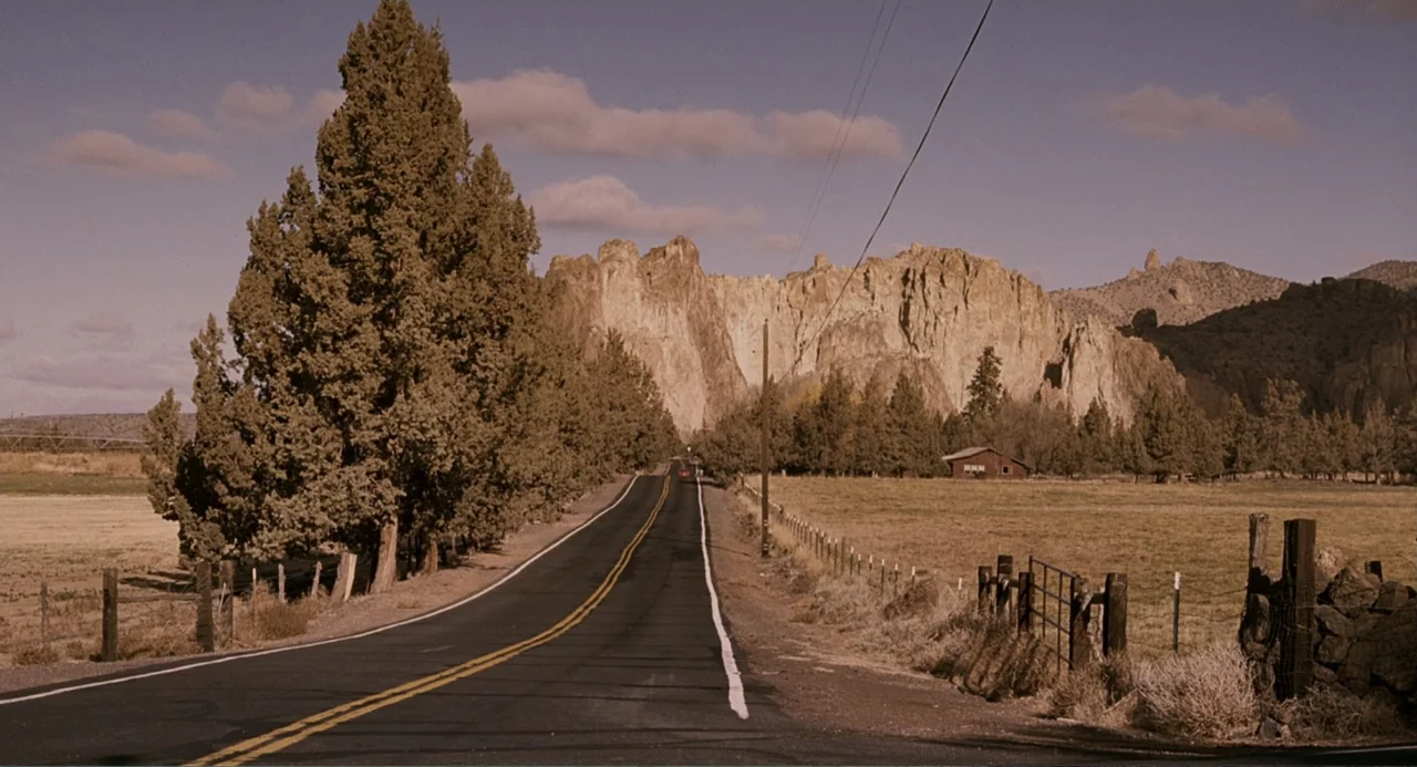 Management (2009) movie still: establishing — A rural asphalt highway winds through a dry landscape with tall conifers and distant cliffs. A small…