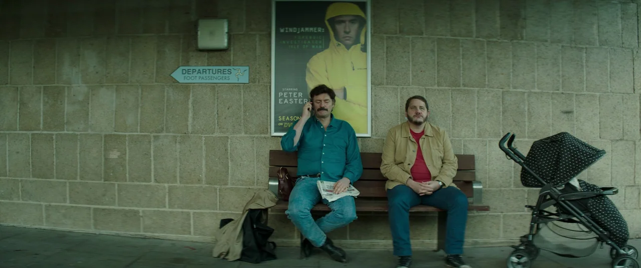 Mindhorn (2016) movie still: wide — Two men wait on a wooden bench before a textured, light-colored stone wall. A sign for ferry departures and a…