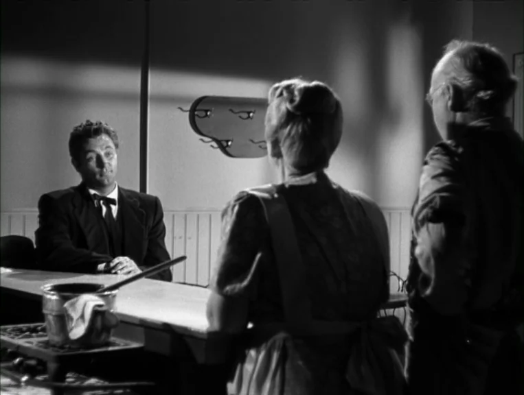 The Night Of The Hunter (1955) movie still: medium — A dimly lit room features a small table and a stylized wall ornament. Three individuals…