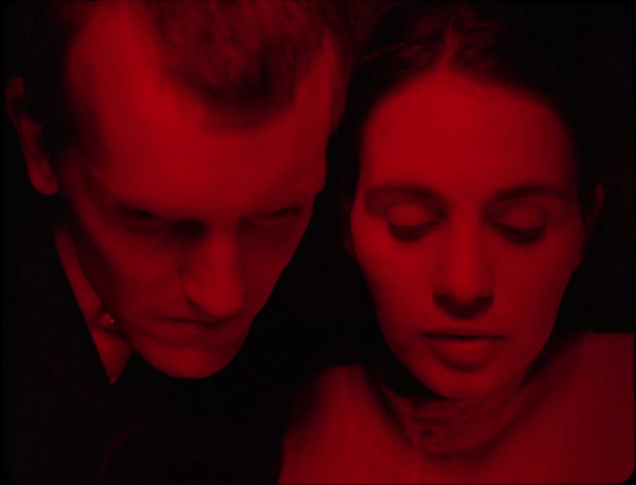 Godland (2022) movie still: closeup — Two individuals stand in an enclosed, shadow-filled environment lit by a single crimson source. They look down…