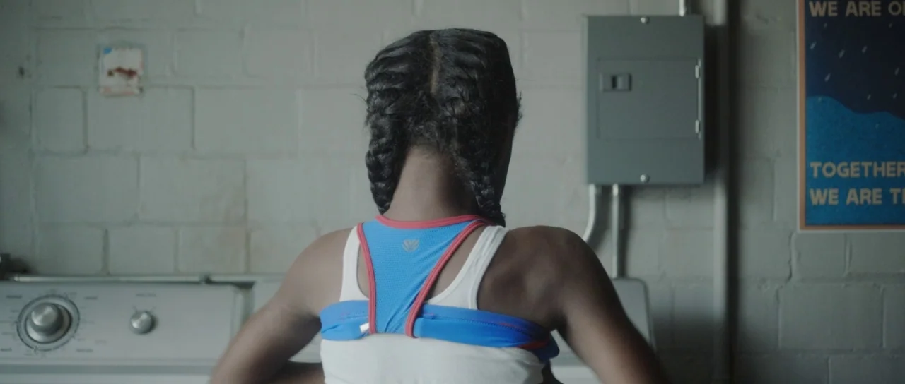 The Fits (2016) movie still: medium — A basement laundry room features gray painted brick walls and a metal electrical box. A girl stands in front…