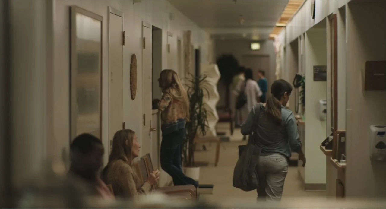 Beatriz at Dinner (2017) movie still: medium — A long interior hallway features rows of chairs and several doors along the walls. A woman with a…