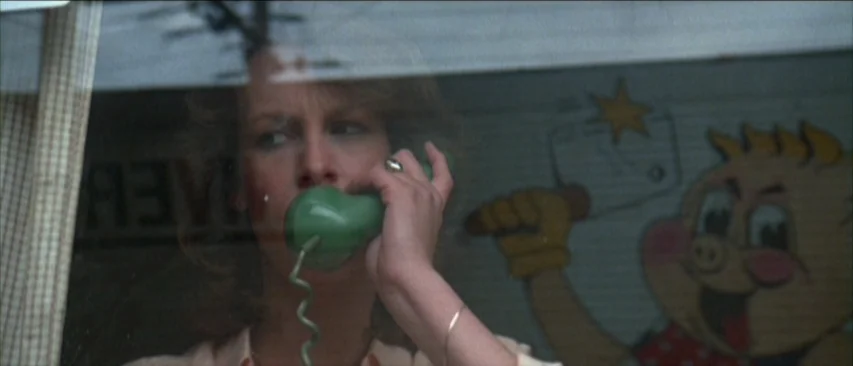 Roadgames (1981) movie still: closeup — A woman looks out through the window of a shop with a pig character decal. She holds a green landline…