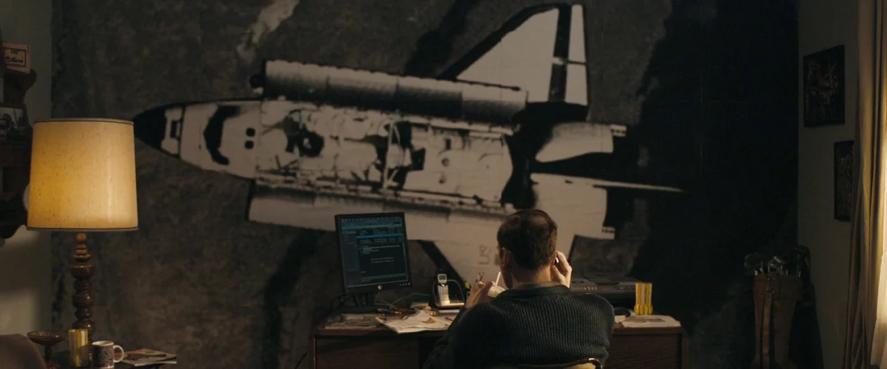 Compliance (2012) movie still: medium — A dark home office features a large black and white space shuttle mural on the wall. A man sits at a wooden…