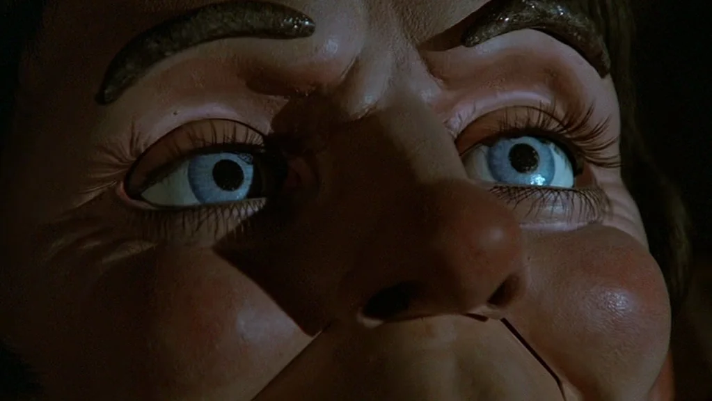 Magic (1978) movie still: closeup — A close-up view of a ventriloquist doll face with blue glass eyes. The doll has a rigid mouth line across the…