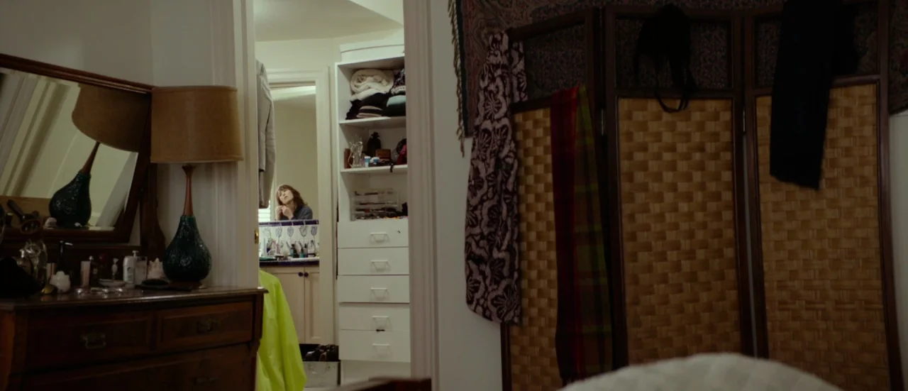 Rupture (2016) movie still: wide — A cluttered residential dressing room with a wooden vanity and a woven room divider. A woman stands in the…