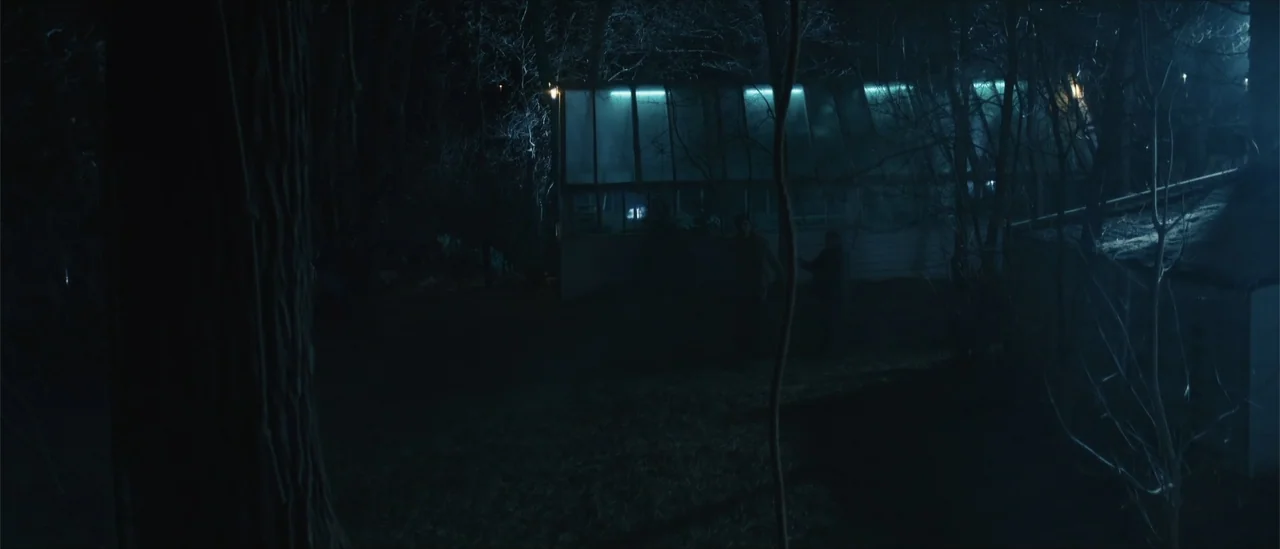 All Fun and Games (2023) movie still: wide — A dark yard features a glowing glass conservatory surrounded by sparse, leafless trees at night. Two…